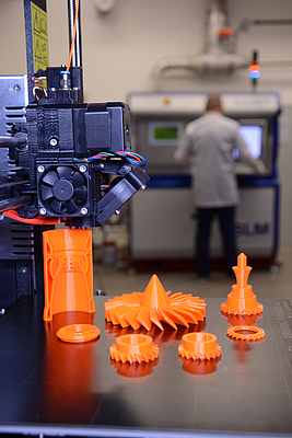 3D-Drucker und 3D-gedruckte Zahnräder, im Hintergrund eine Person an einer Anlage 