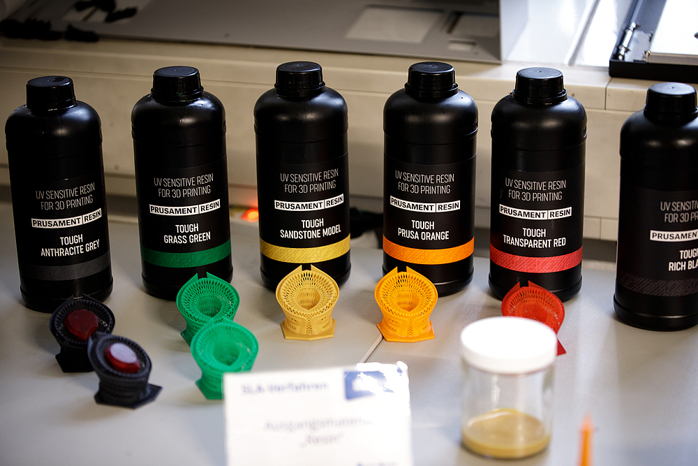 Mehrere Kunststoffflaschen stehen im Halbkreis. Davor liegen 3D Druck Elemente in verschiedenen Farben.