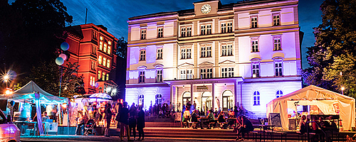 Beleuchtetes Gebäude in der Nacht mit vielen Menschen davor. Fröhliche Stimmung