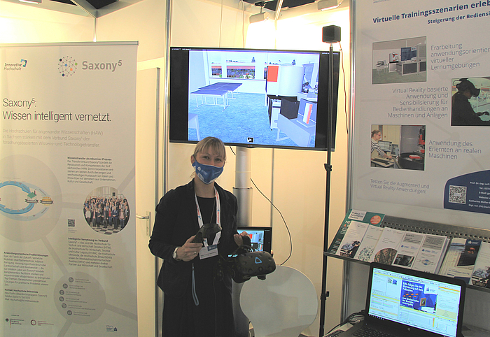 Frau mit Mund-Nasen-Maske steht vor einem Bildschirm und hält eine VR-Brille in der Hand