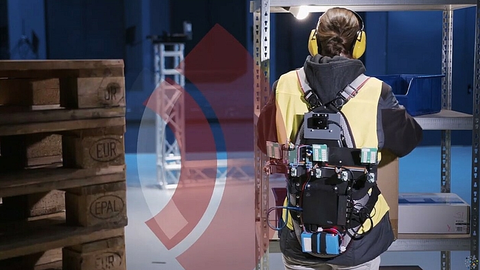 Eine Frau mit gelber Warnweste, bestückt mit Elektronik, arbeitet mit dem Rücken zu einem Palettenstapel, Schallwellen vom Stapel zur Frau sind eingezeichnet